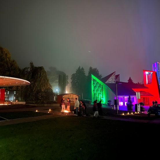 Eine Nachtszene mit einer Gruppe von Menschen, die um ein Lagerfeuer unter einem nebligen Himmel stehen. Der Bereich ist von farbigen Lichtern eines Gebäudes beleuchtet.