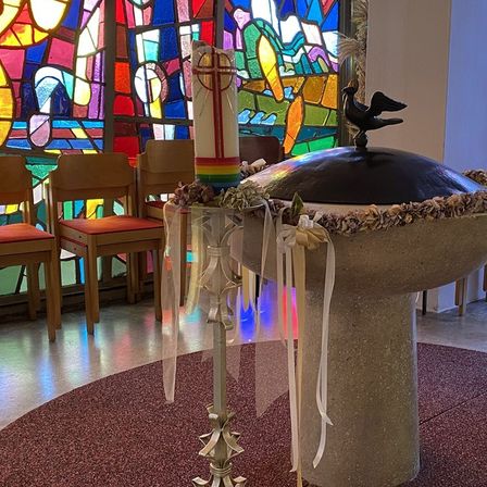 Ein Taufbecken ist mit Muscheln und Bändern verziert vor einem Buntglasfenster in einer Kirche. Stühle sind hinter dem Becken angeordnet.