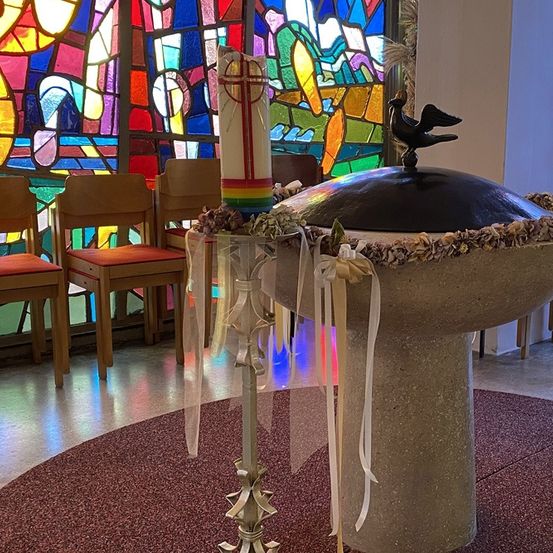 Ein Taufbecken ist mit Muscheln und Bändern verziert vor einem Buntglasfenster in einer Kirche. Stühle sind hinter dem Becken angeordnet.