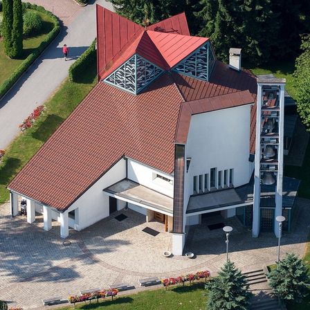 Ein Luftbild einer modernen weißen Kirche mit rotem Dach und Glockenturm. Sie ist von grünem Gras und Bäumen umgeben. Eine Person geht auf dem Weg.
