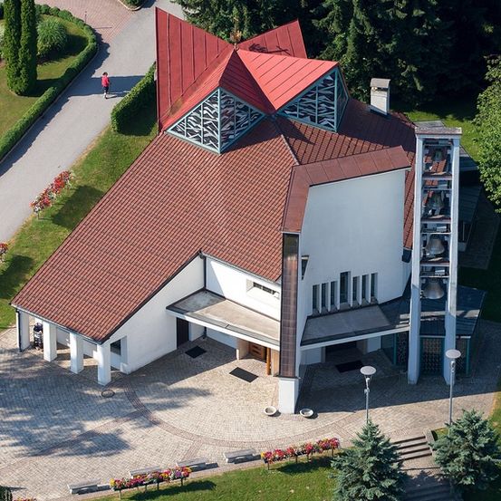 Ein Luftbild einer modernen weißen Kirche mit rotem Dach und Glockenturm. Sie ist von grünem Gras und Bäumen umgeben. Eine Person geht auf dem Weg.