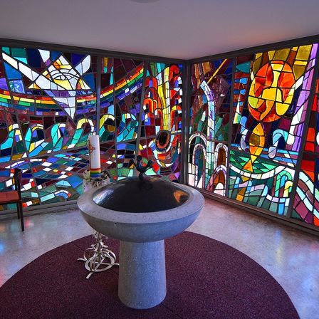 Ein ruhiger Raum mit farbenfrohen Buntglasfenstern zeigt komplizierte Muster. Eine Taufschale steht in der Mitte, mit einer Kerze auf einem Ständer in der Nähe. Ein Stuhl steht links.