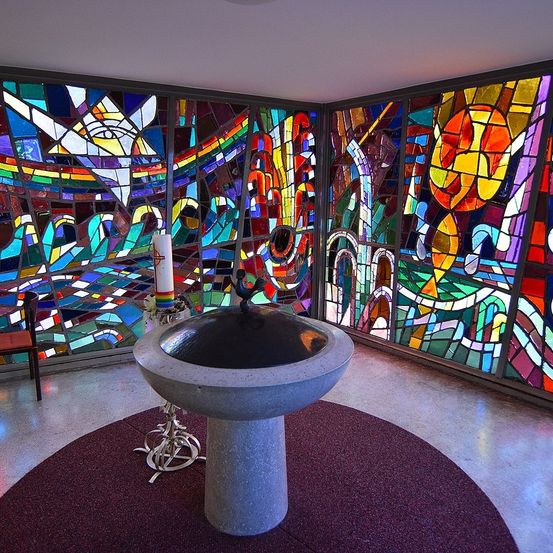 Ein ruhiger Raum mit farbenfrohen Buntglasfenstern zeigt komplizierte Muster. Eine Taufschale steht in der Mitte, mit einer Kerze auf einem Ständer in der Nähe. Ein Stuhl steht links.