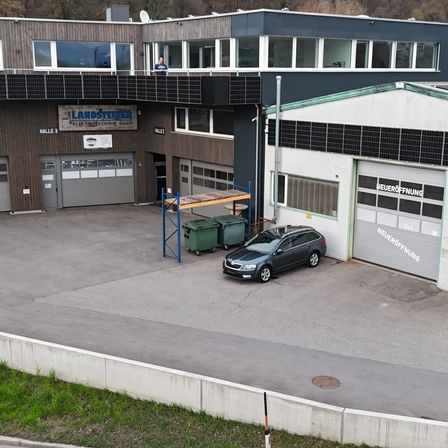 Eine Luftaufnahme eines Industriegebäudes mit Solarmodulen, einem geparkten Auto und zwei Mülleimern. Das Gebäude hat ein Schild mit der Aufschrift 'LANDSTEINER' und ein weiteres Schild mit der Aufschrift 'NEUEROEFFNUNG'.