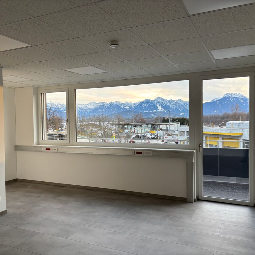 Ein leerer Raum mit weißen Wänden, gefliestem Boden und großen Fenstern mit Blick auf Berge. Es gibt zwei Schalter an der Wand.