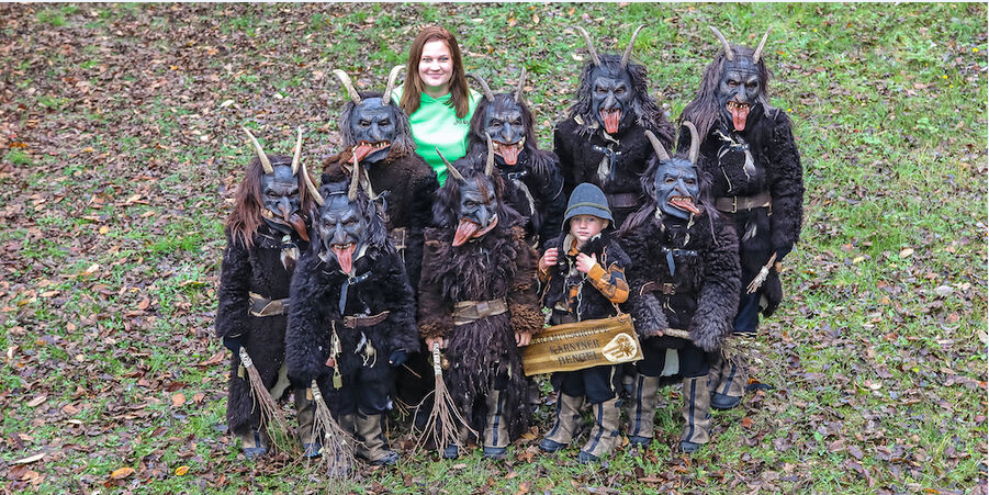Eine Gruppe von Menschen in Hörnerkostümen steht auf einem Rasen. Eine Frau in einem grünen Shirt steht unter ihnen. Ein kleiner Junge hält ein Schild.