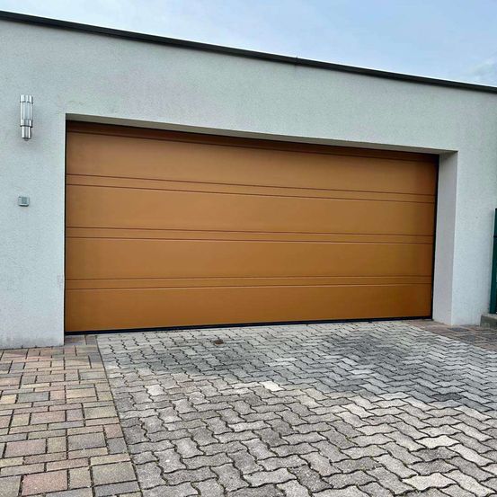 Eine beigefarbene Garagentür mit vertikalen Streifen ist geschlossen und an einer weißen Wand mit einer Lampe montiert. Ein gepflasterter Vorplatz befindet sich vor der Garage.