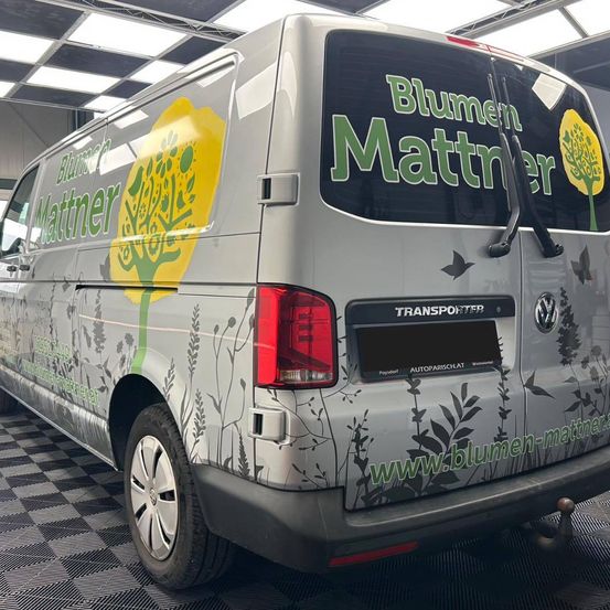 Die Rückseite eines silbernen Volkswagen Transporter-Vans mit grünen und gelben Blumendesigns an der Seite und hinten und dem Text 'Blumen Mattner' auf der Heckscheibe.