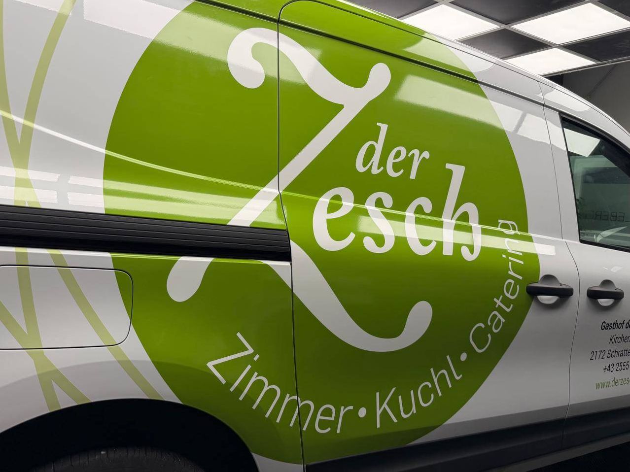 Die Seite eines Lieferwagens zeigt einen grünen Kreis mit weißen Buchstaben, auf dem 'Zimmer Kuchl Catering' steht. Der Kreis enthält das Logo 'der Zesch'. Der Lieferwagen ist weiß mit grünen Akzenten lackiert.