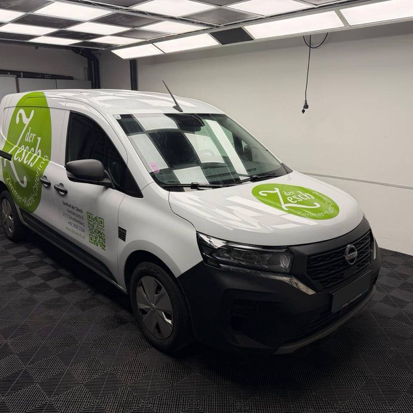 Ein weißer Lieferwagen mit grüner Beschriftung steht in einer Garage mit einem karierten Boden. Er trägt ein Logo mit dem Text 'Zesch' und einer Telefonnummer.