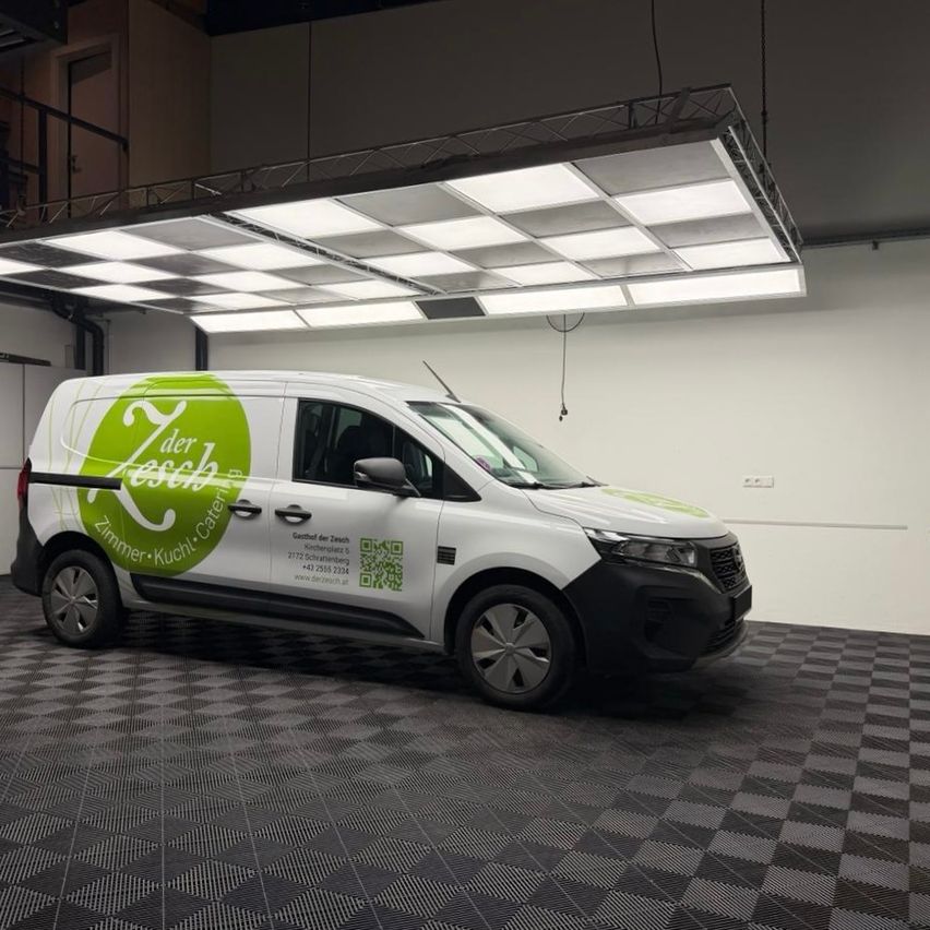 Ein weißer Van mit grün-weißer Beklebung steht in einem Raum mit gemustertem Boden und großen Leuchten darüber. Der Van ist mit einem Logo und Text beklebt.