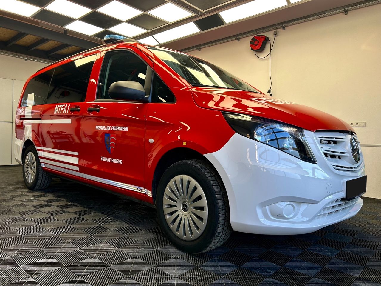 Ein rot-weißer Mercedes-Benz-Fahrzeug mit 'Feuerwehr'-Kennzeichen steht in einer Garage. Das Fahrzeug hat die Aufschrift 'MTFA1' an der Tür und eine rote Sirene auf dem Dach.