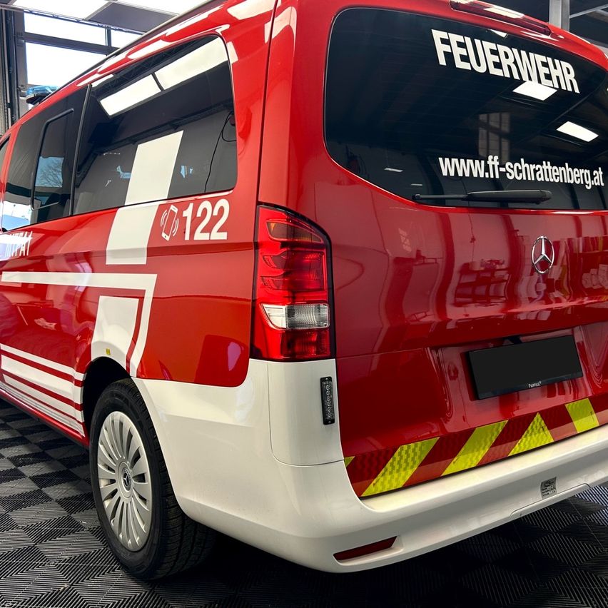 Ein rotes Fahrzeug mit weißen und roten Streifen, mit einer Website-Adresse auf der Heckscheibe und einer Telefonnummer an der Seite. Das Fahrzeug steht auf einem gemusterten Boden.