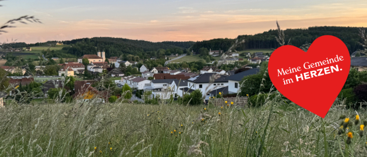 Ein Dorf in einem Tal mit einem Sonnenuntergang im Hintergrund. Ein rotes Herz mit der Aufschrift 'Meine Gemeinde im Herzen' ist im Vordergrund.