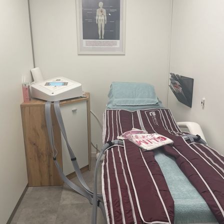 Ein Behandlungsraum mit einem Bett, medizinischen Geräten und einem Diagramm an der Wand. Das Bett hat eine marmorierte und weiße gestreifte Decke.
