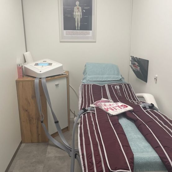 Ein Behandlungsraum mit einem Bett, medizinischen Geräten und einem Diagramm an der Wand. Das Bett hat eine marmorierte und weiße gestreifte Decke.