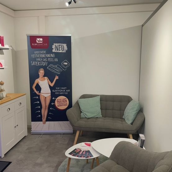 Ein Raum mit einer grauen Couch, einem weißen Tisch, einem Schrank und einem Roll-up-Banner mit einem Bild einer Frau und Text über effektive Fettverbrennung.