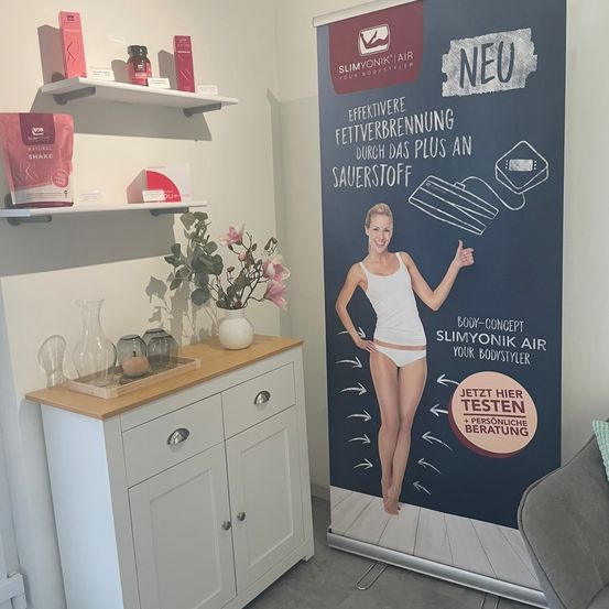 Ein weißer Schrank mit Schubladen steht neben einem Plakat mit einer Frau, einer Blumenvase und einem roten Pulverbeutel auf den Regalen. Rechts ist eine Couch.