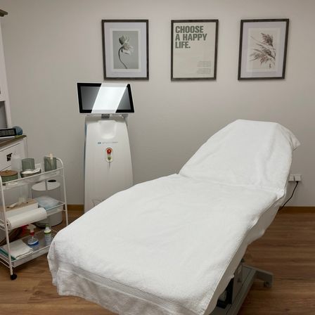 Ein medizinischer Raum mit einem weißen Bett und einem Monitor, begleitet von einem kleinen Tisch und gerahmten Bildern an der Wand.