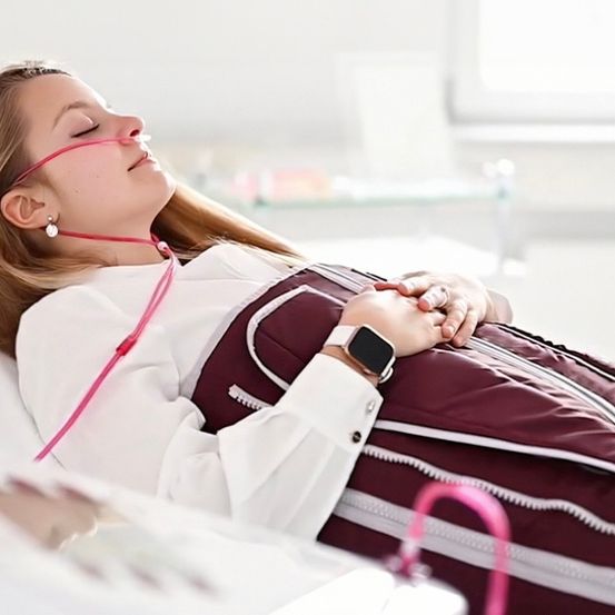 Eine Frau liegt mit einer Maske über dem Gesicht auf einem Krankenhausbett, trägt ein weißes Hemd und eine kastanienbraune Decke. Sie hält eine Hand auf die Brust.