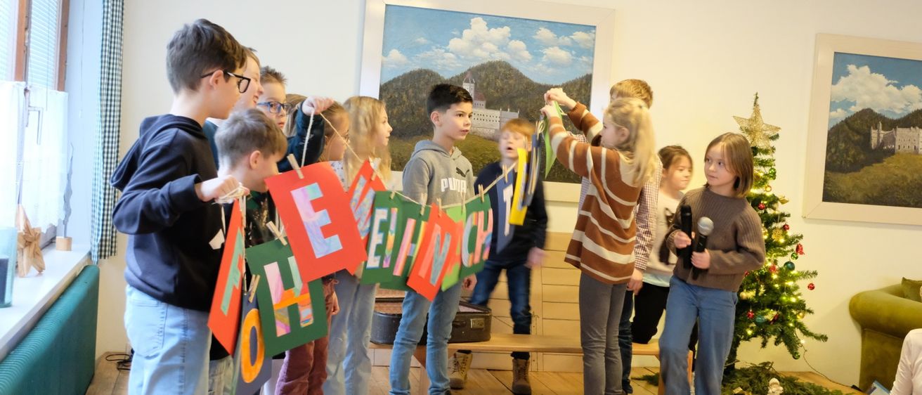 Eine Gruppe von Kindern steht in einem Raum mit einem Weihnachtsbaum und hält bunte Banner, die eine Kette bilden. Ein Mädchen in einem gestreiften Shirt spricht in ein Mikrofon. Hinter ihnen ist ein Gemälde von Bergen und einem Schloss.