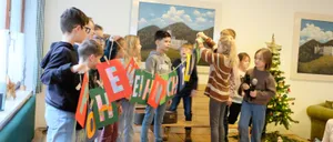 Eine Gruppe von Kindern steht in einem Raum mit einem Weihnachtsbaum und hält bunte Banner, die eine Kette bilden. Ein Mädchen in einem gestreiften Shirt spricht in ein Mikrofon. Hinter ihnen ist ein Gemälde von Bergen und einem Schloss.