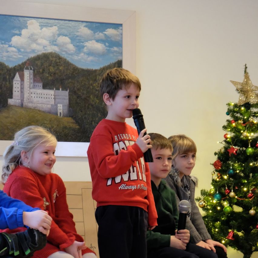 Ein kleiner Junge in einem roten Sweatshirt spricht in ein Mikrofon, während er mit anderen Kindern vor einem Weihnachtsbaum und einem Bild eines Schlosses sitzt.