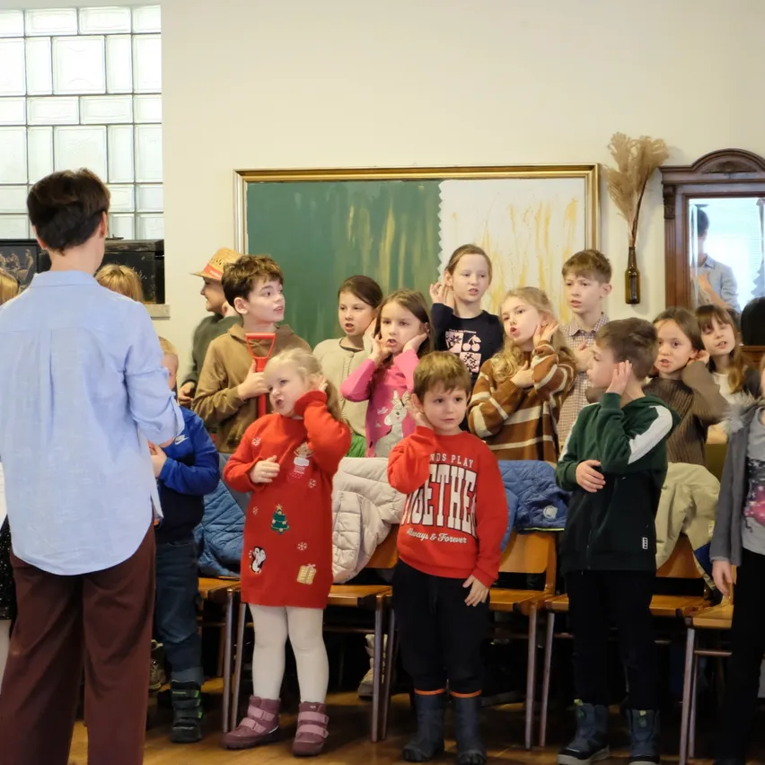 Eine Gruppe von Kindern und einem Mann stehen in einem Raum. Einige Kinder tragen Stiefel und andere Mützen. Ein Mann trägt ein blaues Hemd und braune Hosen. Ein Gemälde und ein Spiegel sind an der Wand.