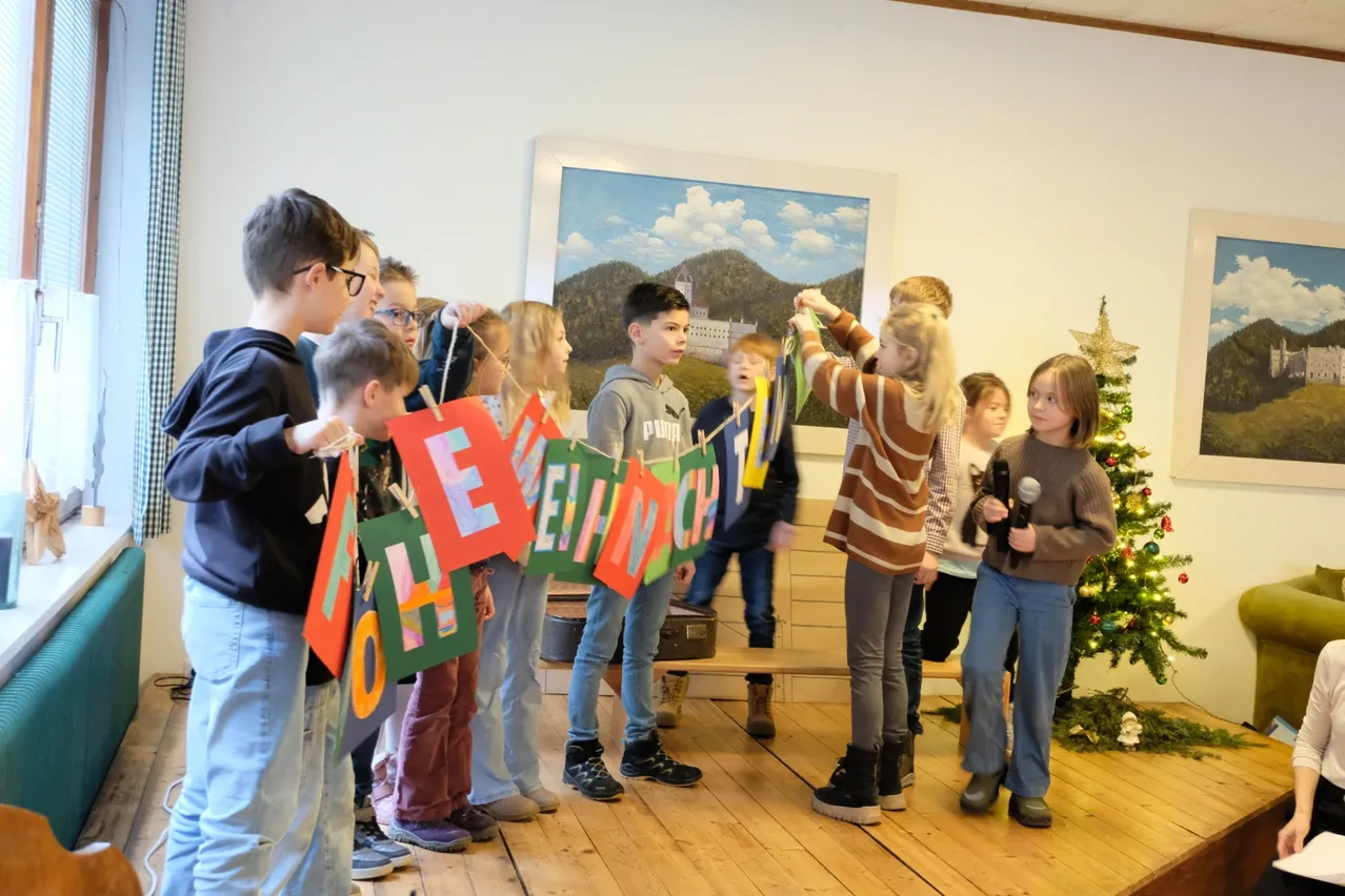 Eine Gruppe von Kindern hält bunte Banner mit dem Wort 'Willkommen' in einem Raum hoch. Ein Mädchen hält ein Mikrofon und ein anderes Mädchen hebt die Hände. Ein Weihnachtsbaum steht rechts. Hinter ihnen ist eine Bank mit einem Koffer darauf. Ein Gemälde ist an der Wand.
