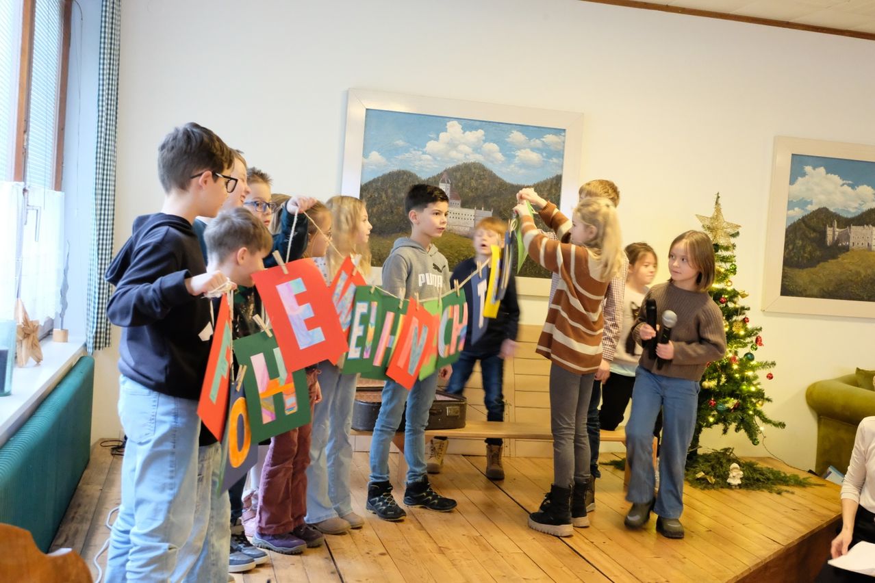 Eine Gruppe von Kindern hält bunte Banner mit dem Wort 'Willkommen' in einem Raum hoch. Ein Mädchen hält ein Mikrofon und ein anderes Mädchen hebt die Hände. Ein Weihnachtsbaum steht rechts. Hinter ihnen ist eine Bank mit einem Koffer darauf. Ein Gemälde ist an der Wand.