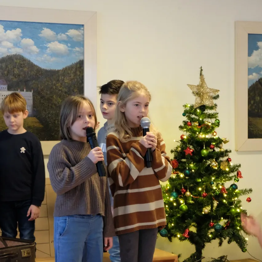 Vier Kinder stehen vor einem Weihnachtsbaum, jedes hält ein Mikrofon in der Hand. Die Kinder sind in Winterkleidung gekleidet. Hinter ihnen hängen zwei gerahmte Gemälde an der Wand.