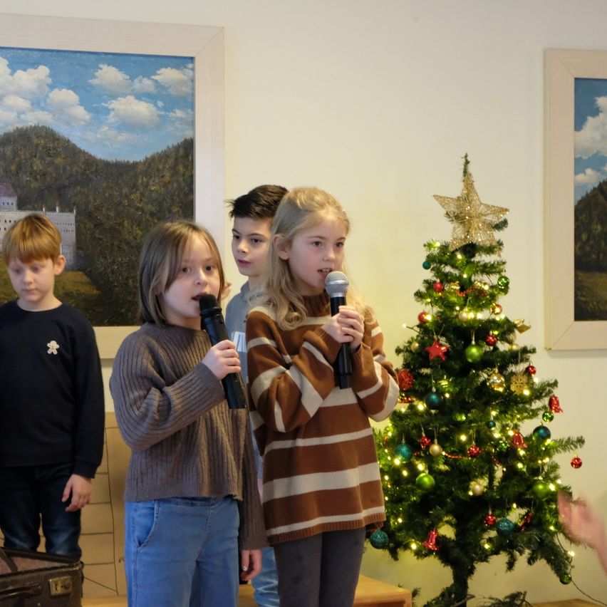 Vier Kinder stehen vor einem Weihnachtsbaum, jedes hält ein Mikrofon in der Hand. Die Kinder sind in Winterkleidung gekleidet. Hinter ihnen hängen zwei gerahmte Gemälde an der Wand.