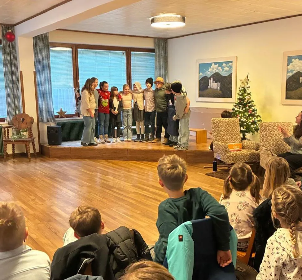 Mehrere Kinder stehen auf einer Bühne in einem Raum, tragen Hüte und Stiefel. Sie scheinen für ein Publikum aufzutreten. Eine Weihnachtsbaum steht in der Ecke.