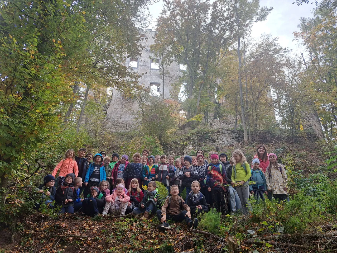 Eine Gruppe von Kindern und Erwachsenen versammelt sich im Wald und posiert für ein Foto mit einem zerstörten Gebäude im Hintergrund.