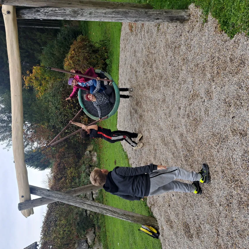 Eine Gruppe von Kindern genießt eine Schaukel auf einem Spielplatz, wobei ein Kind in der Schaukel sitzt und andere die Griffe halten. Der Spielplatz hat eine Kiesoberfläche.