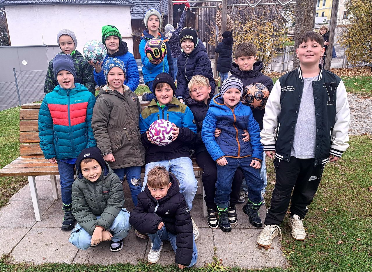 A group of children in winter clothing, posing for a photo with soccer balls. Some hold the balls while others stand behind them, all smiling. They are on a concrete sidewalk with a wooden bench in the background.