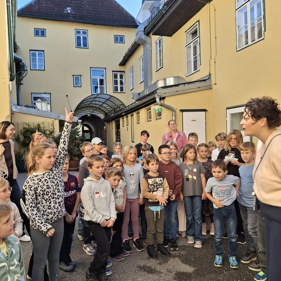 Eine Gruppe von Kindern und Erwachsenen versammelt sich vor einem gelben Gebäude, möglicherweise für eine Schulveranstaltung. Eine Frau spricht zu ihnen.