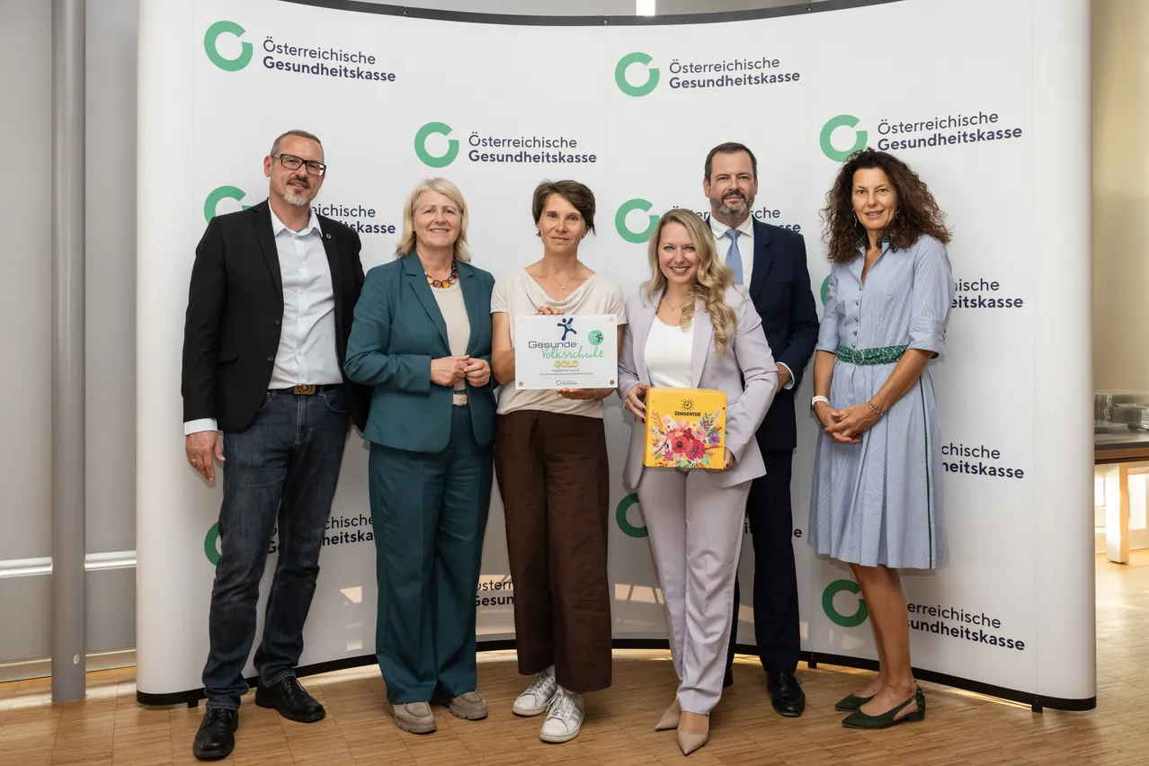 Eine Gruppe von sechs Personen steht zusammen vor einem weißen Banner mit grünem Text und Logos. Sie alle lächeln und scheinen bei einer Preisverleihung zu sein.
