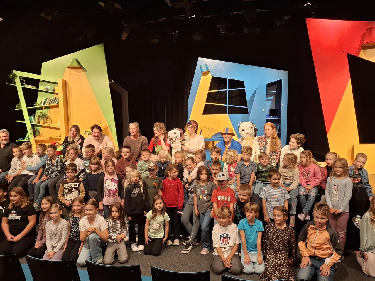 Eine Gruppe von Kindern und Erwachsenen posiert für ein Foto auf einer Bühne mit farbenfrohen Kulissen.
