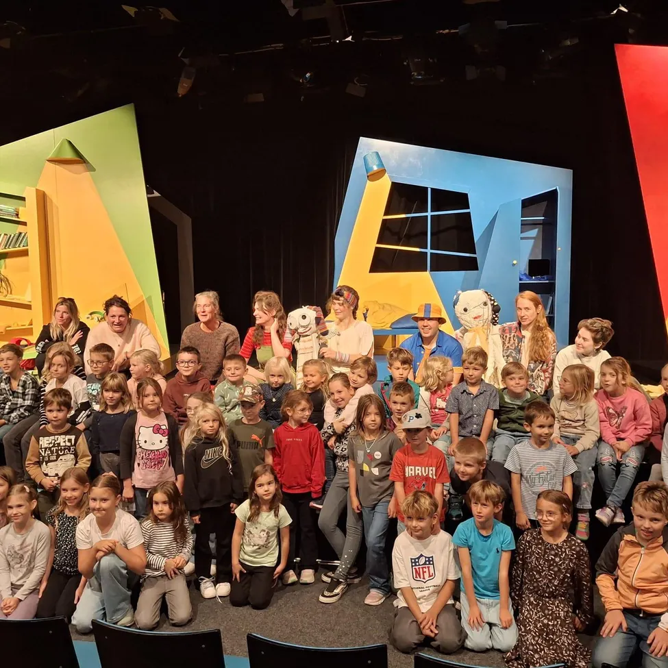 Eine Gruppe von Kindern und Erwachsenen posiert für ein Foto auf einer Bühne mit farbenfrohen Kulissen.