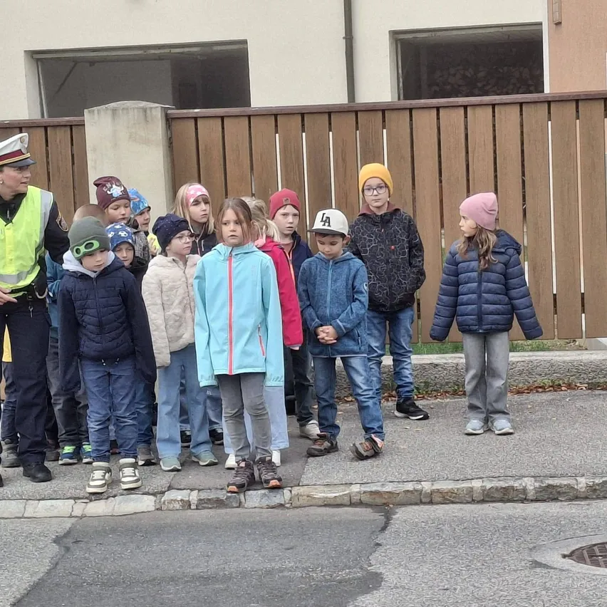 Eine Gruppe von Kindern steht auf dem Bürgersteig, mit zwei Polizeibeamten auf beiden Seiten der Gruppe. Die Kinder tragen Winterkleidung, und einige tragen Mützen und Brillen.