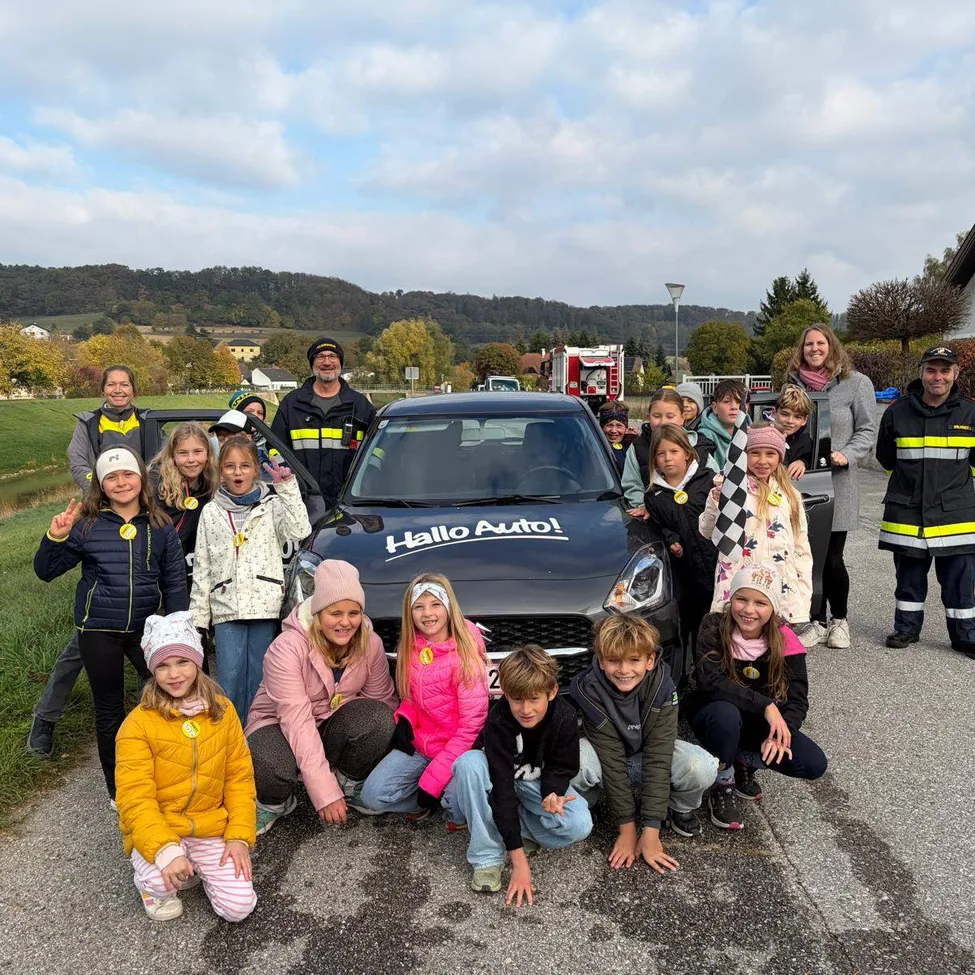 Eine Gruppe von Kindern und Erwachsenen posiert für ein Foto vor einem Auto mit der Aufschrift 'Hallo Auto!' auf der Motorhaube.
