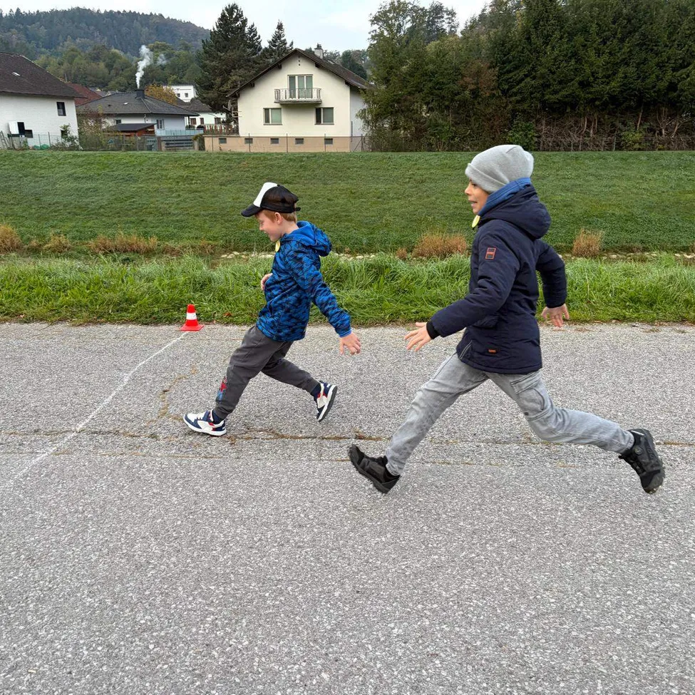 Zwei Jungen rennen auf einer Straße mit einem Grasfeld im Hintergrund. Ein Junge trägt einen blauen Mantel und der andere einen schwarzen Mantel.