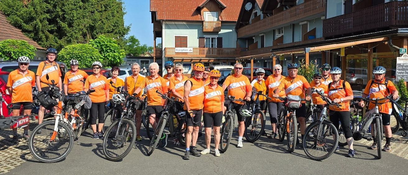 Eine Gruppe von Menschen in orangefarbenen Westen und Helmen steht mit ihren Fahrrädern vor einem Gebäude.