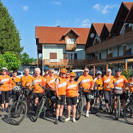 Eine Gruppe von Menschen in orangefarbenen Fahrradtrikots und Helmen steht vor einem Gebäude. Sie posieren für ein Foto.