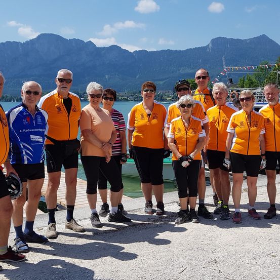 Eine Gruppe von Radfahrern in passenden orangefarbenen Shirts und schwarzen Shorts steht auf einem Steg am See mit Bergen im Hintergrund.