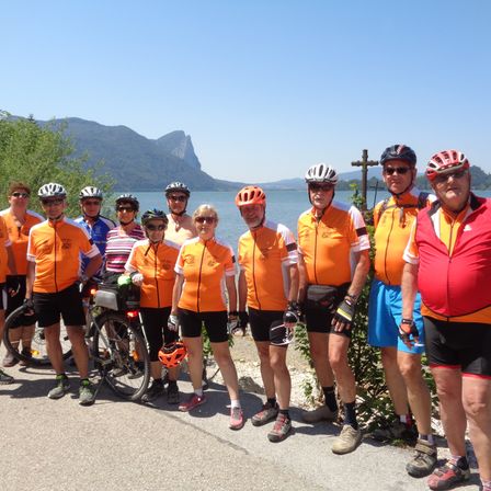 Eine Gruppe von Radfahrern in orangefarbenen Trikots steht am Ufer eines Sees, mit Bergen im Hintergrund. Sie tragen alle Helme und Sonnenbrillen. Einige haben Fahrräder neben sich.