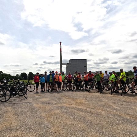 Eine Gruppe von Radfahrern in Helmen und Radbekleidung posiert für ein Foto auf einem Schotterweg. Dahinter stehen Fahrräder und ein Gebäude mit Schornstein. Der Himmel ist bewölkt.