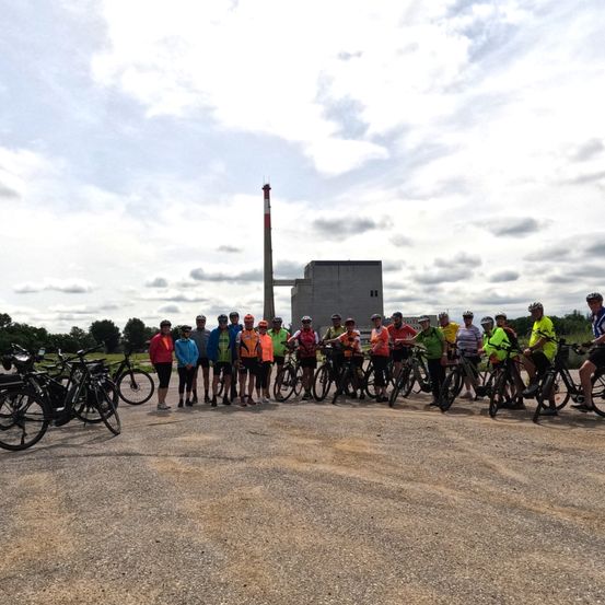 Eine Gruppe von Radfahrern in Helmen und Radbekleidung posiert für ein Foto auf einem Schotterweg. Dahinter stehen Fahrräder und ein Gebäude mit Schornstein. Der Himmel ist bewölkt.