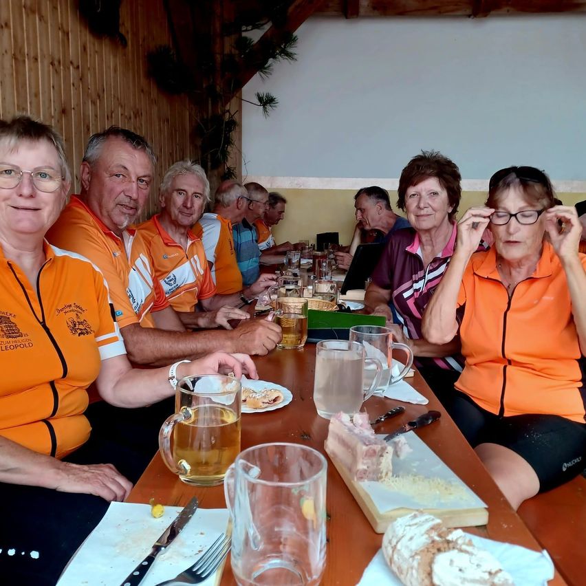 A group of people in orange shirts are sitting at a table, smiling and posing for a photo. They have food, drinks, and glasses on the table.